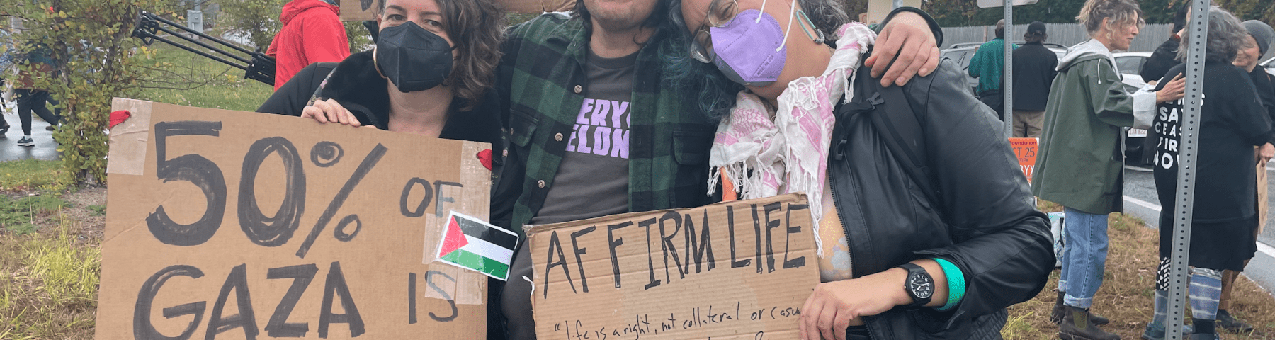 Image of Leah and two friends at a protest. One friend is a white, Greek/Irish femme person in her 40s with greying undercut brown hair wearing a black KN95 mask, they are holding a sign that says 50% OF GAZA IS UNDER 18 STOP KILLING KIDS. The second person is a Syrian masculine person wearing a green hat, a green and black flannel over a black t shirt with lilac lettering, holding a sign that reads CEASEFIRE NOW: FREE PALESTINE with two red black green and white Palestinian flags. Leah stands to the right leaning on their friend with their head on their shoulder, wearing a black leather jacket, black leather skirt, pink and white keffiyeh and lilac KN95 mask, holding a sign that reads, "Affirm life." Protestors can be seen in the background at a rotary in mid fall by a tree