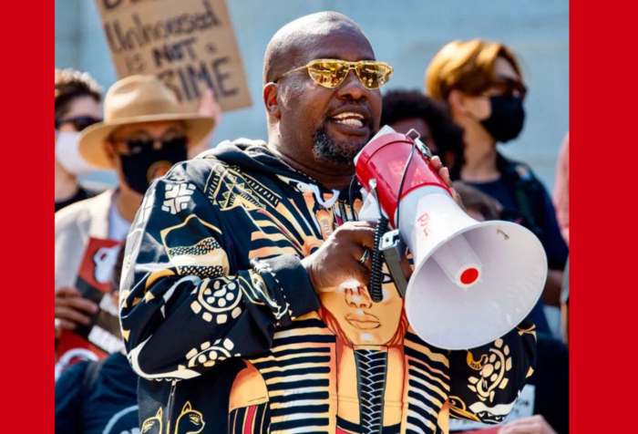 Graphic with a red background and a photo of Theo Henderson, a Black man with a shaved head, goatee, gold-tinted sunglasses, and wearing a hoodie with an image of Tutankhamun on it, speaks into a red and white megaphone at a protest. Behind Theo is a crowd of people wearing face masks, with one holding up a sign that says "Being unhoused is not a crime." Credit: WeTheUnhoused.com