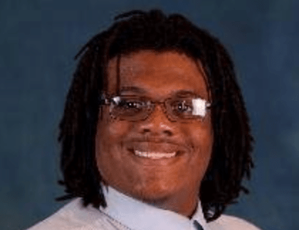 Smiling cis-male with brown complexion, glasses, dark brown eyes, and locs. He is wearing a light blue shirt