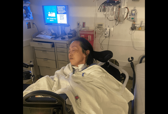 A picture of me, an Asian American disabled woman in a wheelchair, a tracheostomy is in my throat connected to a ventilator tube. I am in the emergency room at UCSF Moffitt/Long hospital. I am swaddled in blankets, my eyes are closed, and I am trying to sleep as I am waiting for test results and a bed in the intensive care unit. Behind me is a computer monitor with a bright glow. On a table in front of me is a wheelchair battery charger.