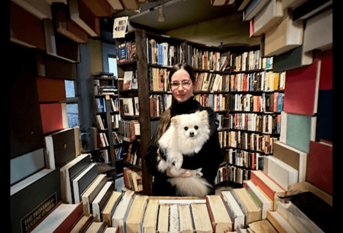 A white woman wearing glasses and a black sweater smiles while holding a small, fluffy white dog in her arms. They are surrounded by tall shelves filled with books, and are gazing together at the camera through a circular “window” opening in a sculptural wall made of hardcover books in an array of colors.
